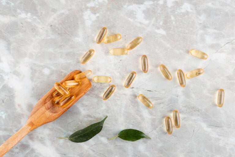 Cápsulas de omega 3 sobre mármol claro, algunas en una cuchara de madera, junto a dos hojas verdes
