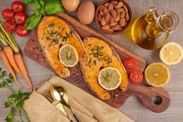 Dos filetes de salmón al horno con limón sobre una tabla, junto a una botella de aceite, almendras, huevos, tomates y zanahorias